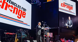 Man speaks at podium at Vehicles for Change event with event logo on screens behind him.