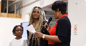 Woman in red speaking into microphone while holding paper, with young girl and woman with braided hair nearby.