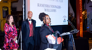 Woman at a podium speaking during an award ceremony; two people stand behind her.