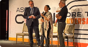 Three people on stage speaking at a conference, one holding a microphone and tablet.