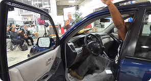 Man sitting in driver seat of blue car raising right arm in an indoor showroom with seated audience.