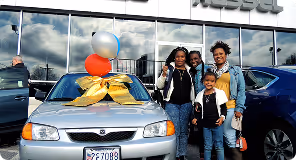 Family of four smiling next to silver car decorated with balloons and a gold ribbon at dealership.