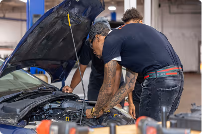 Mechanic in green hoodie working on a car engine with hood open in a garage.