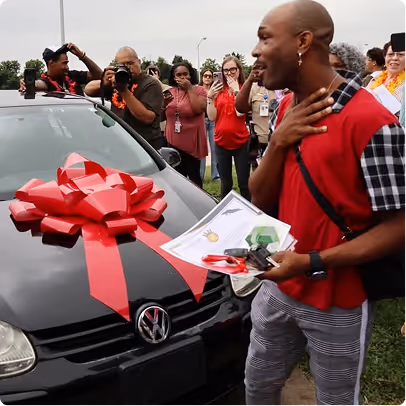Man surprised by a new Volkswagen car with a red bow, holding keys and a certificate, surrounded by applauding people.