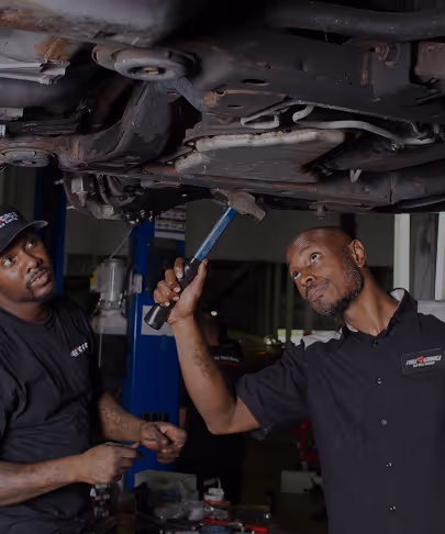 Two mechanics inspecting and working on the underside of a lifted vehicle in a garage.