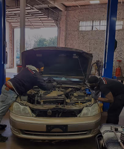 Two mechanics working on an open engine of a beige car in a garage.