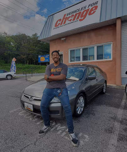 Man smiling and sitting on the hood of a gray Honda parked outside Vehicles for Change building.