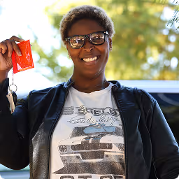 Smiling person wearing glasses and a Shelby Cobra shirt, holding a small red packet and keys outdoors.