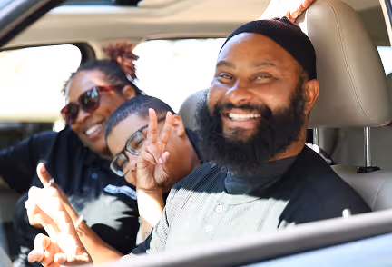 Three smiling people giving peace signs inside a car during daytime.