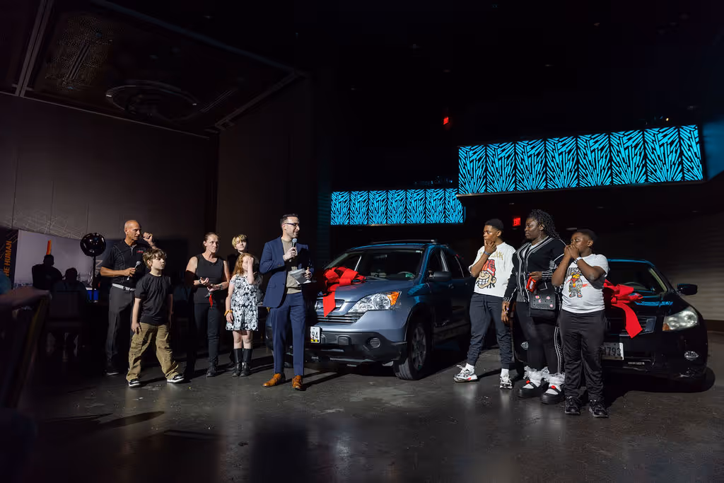 Group of people standing beside two cars with red bows at an indoor event with blue lit decor.