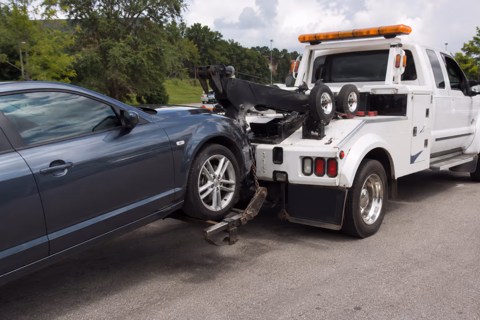 Dark gray car being towed by a white tow truck on a road near green trees.