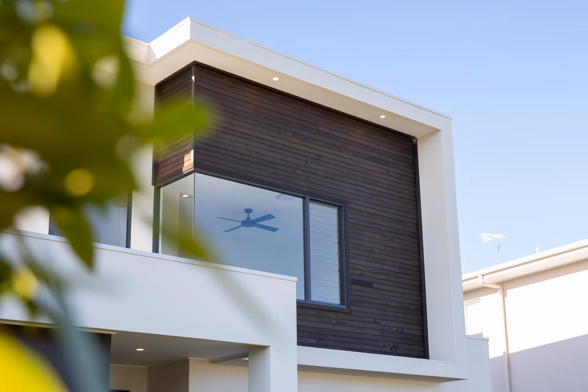 Modern two-story house with white exterior and dark wooden paneling around a large window showing a ceiling fan inside.