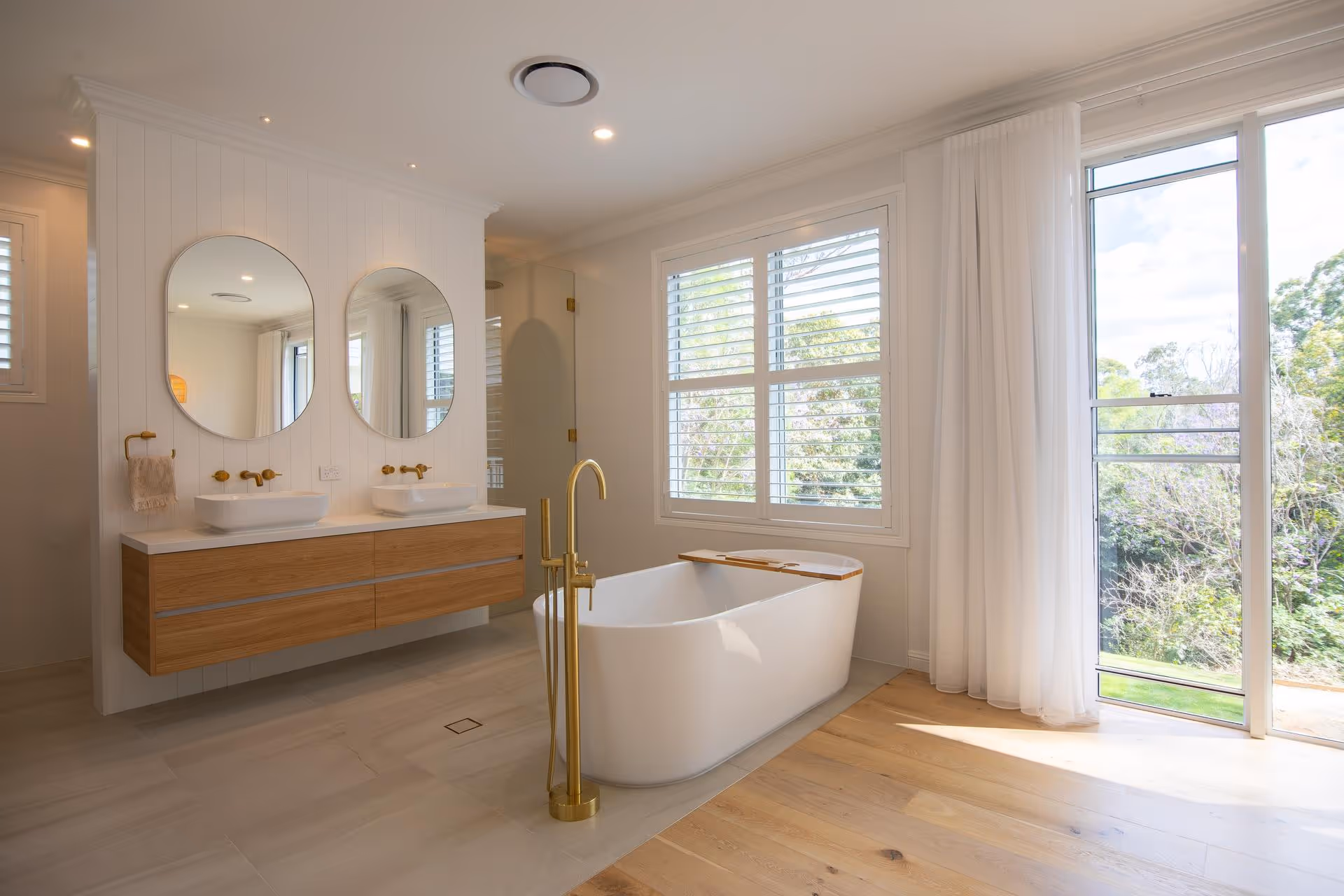 Modern bathroom with freestanding white bathtub, wooden vanity with two oval mirrors, gold fixtures, and large window with white blinds and curtains.