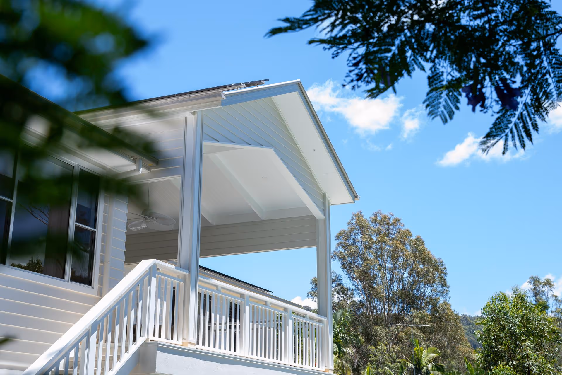 White porch with railings and ceiling fan attached to a house under a blue sky surrounded by trees.