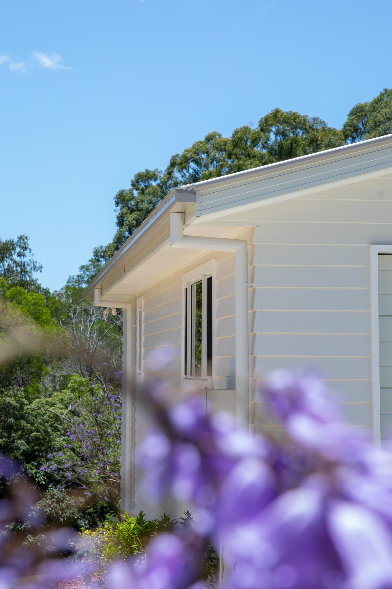Side view of a white house with a small window, surrounded by green trees and purple flowers in the foreground under a clear blue sky.