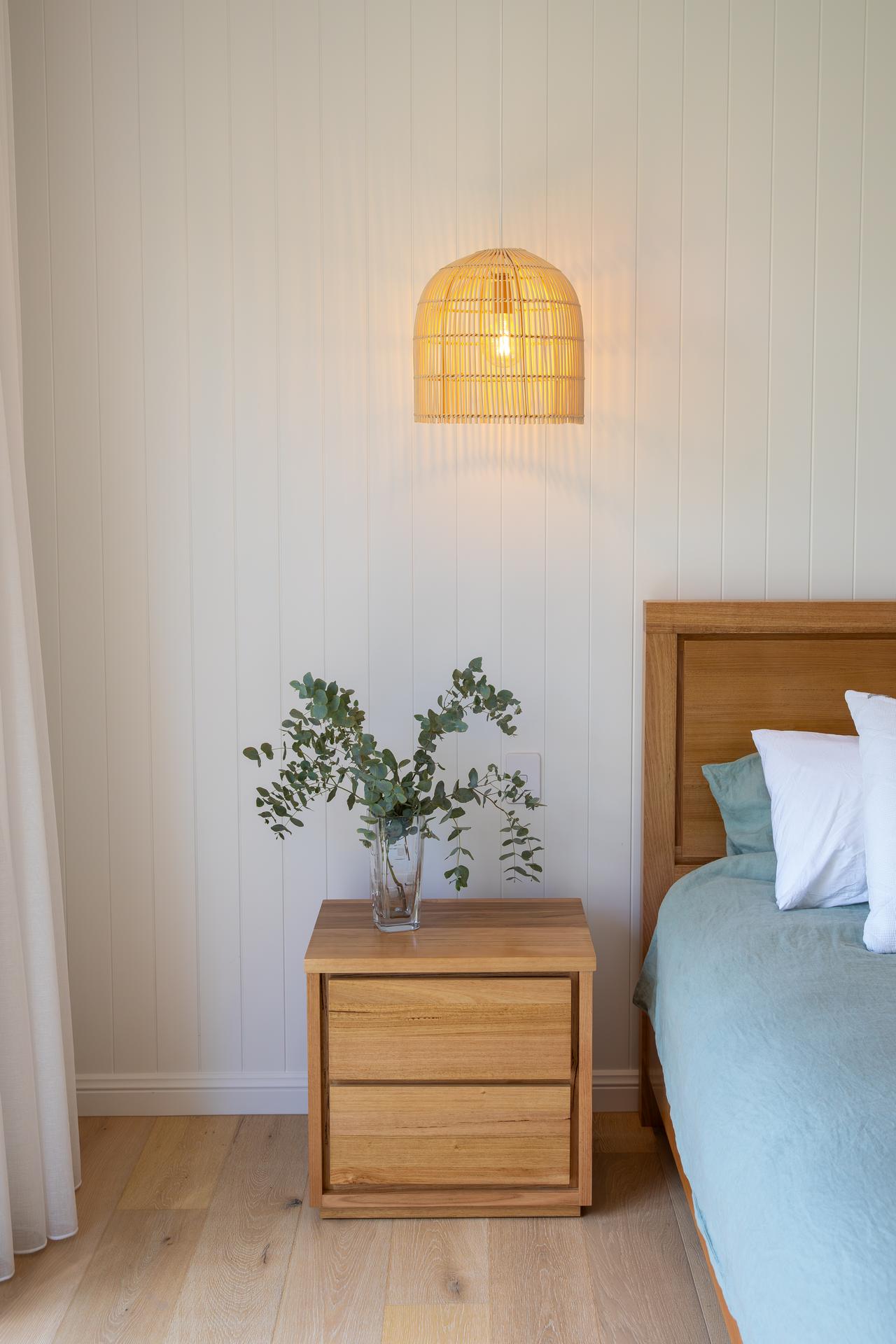Wooden nightstand with a glass vase of eucalyptus and a lit wicker wall lamp in a bedroom.