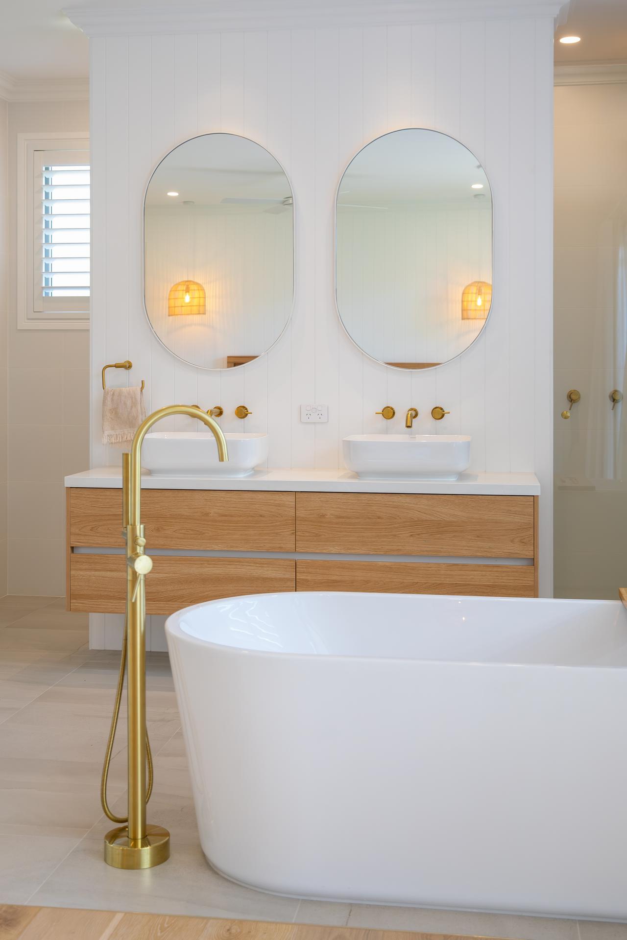 Modern bathroom with freestanding white tub and double sinks with oval mirrors and brass fixtures.