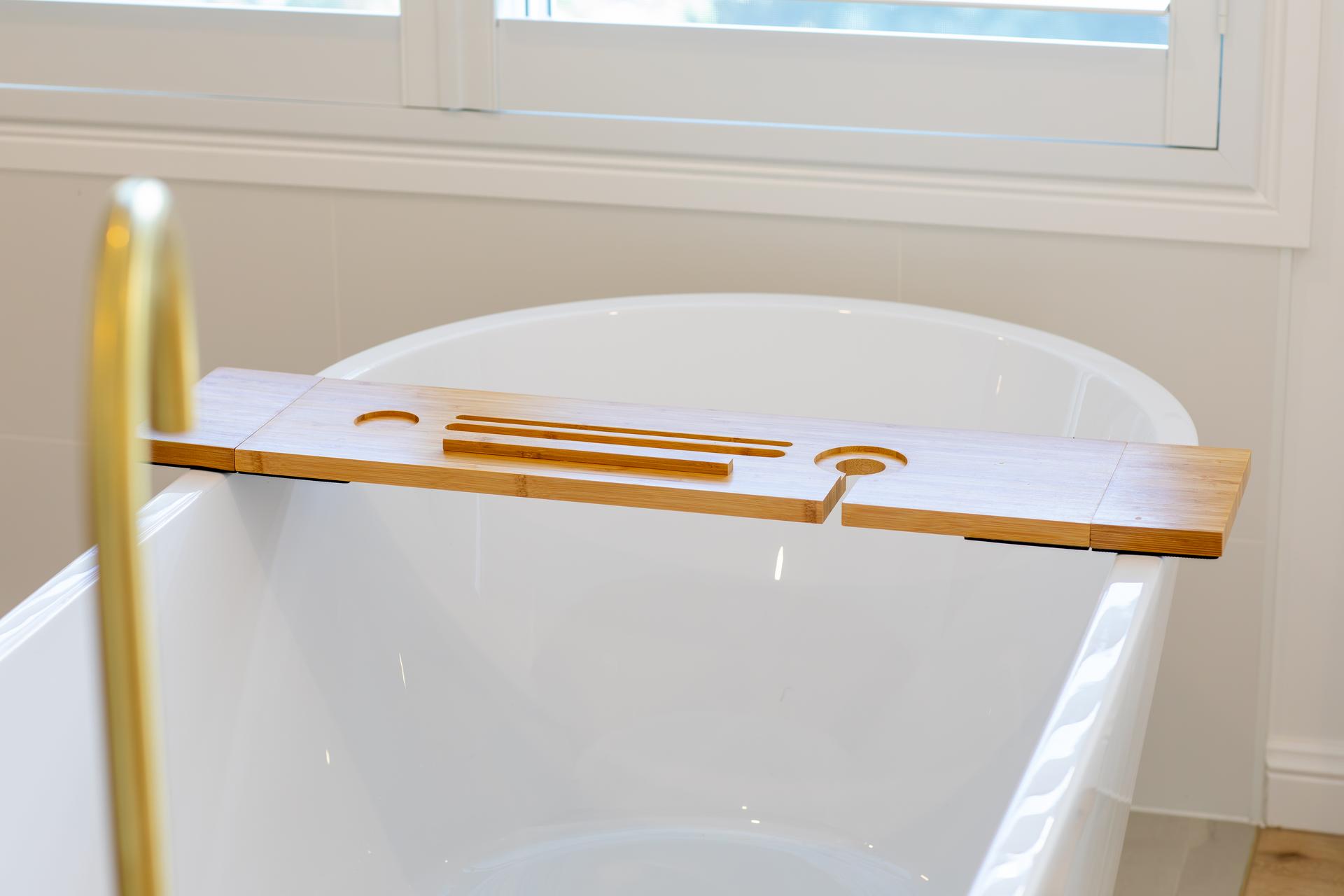 Bamboo bath tray placed over a white freestanding bathtub under a window.