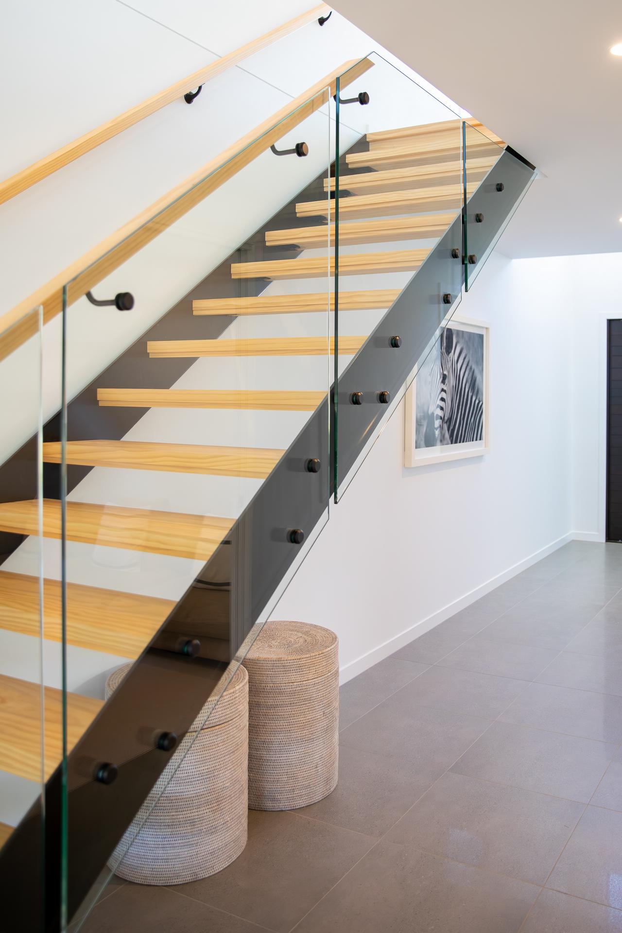 Modern staircase with wooden steps and glass railing over two woven round stools in bright hall.