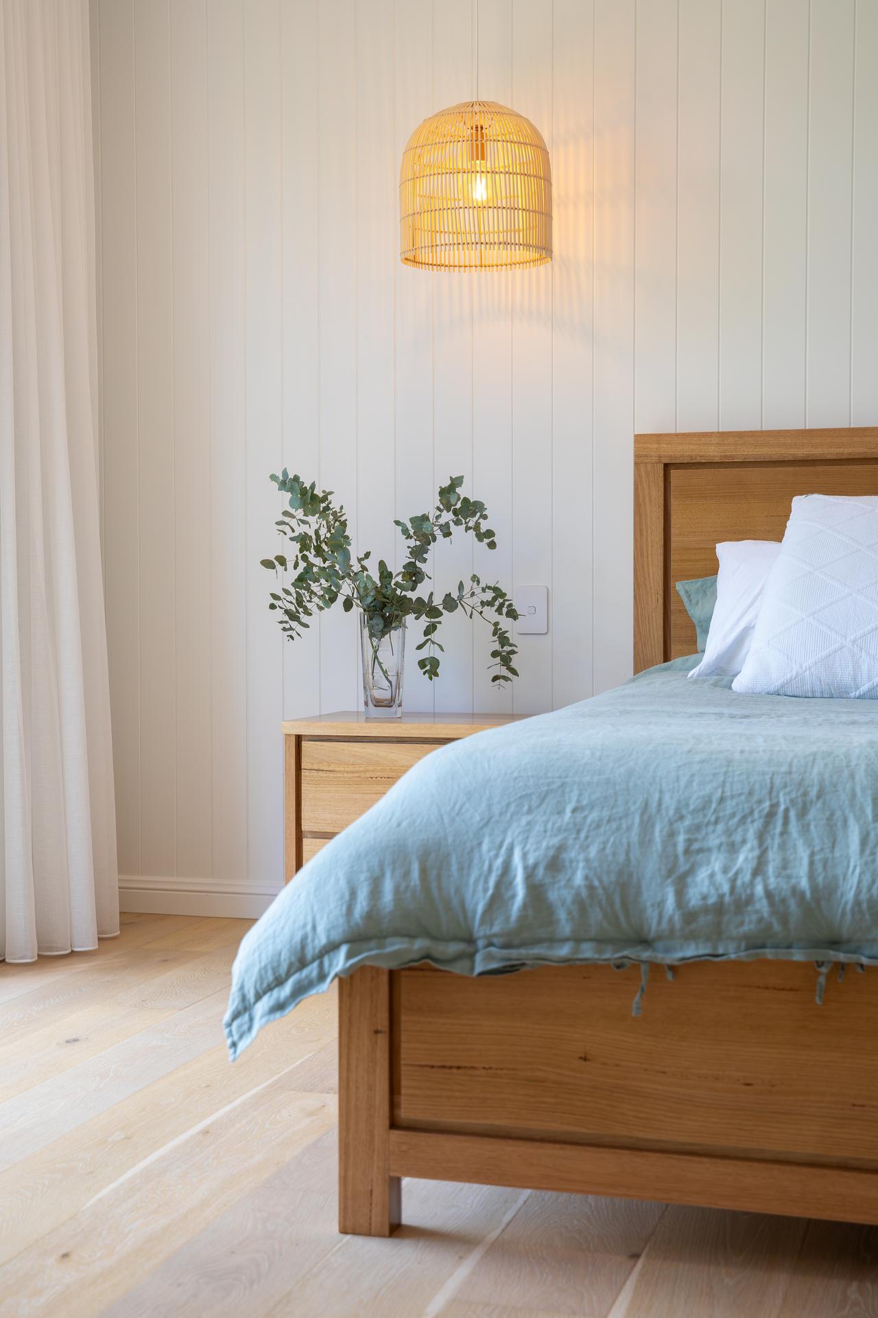 Bedroom corner with wooden bed, blue bedding, white pillows, nightstand, vase with greenery, and hanging lamp.