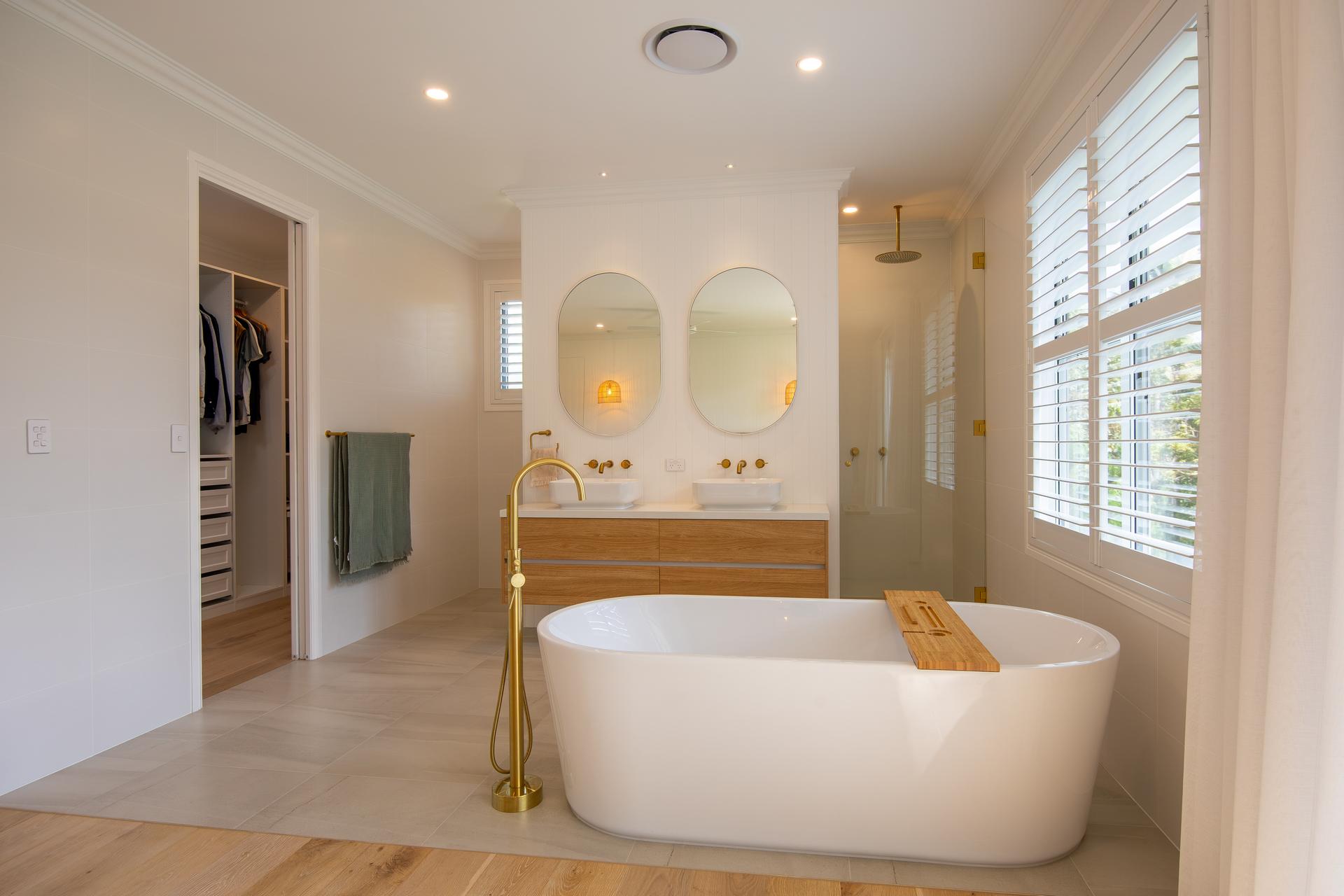Modern bathroom with freestanding tub, wooden vanity with dual sinks, brass fixtures, and large shuttered window.