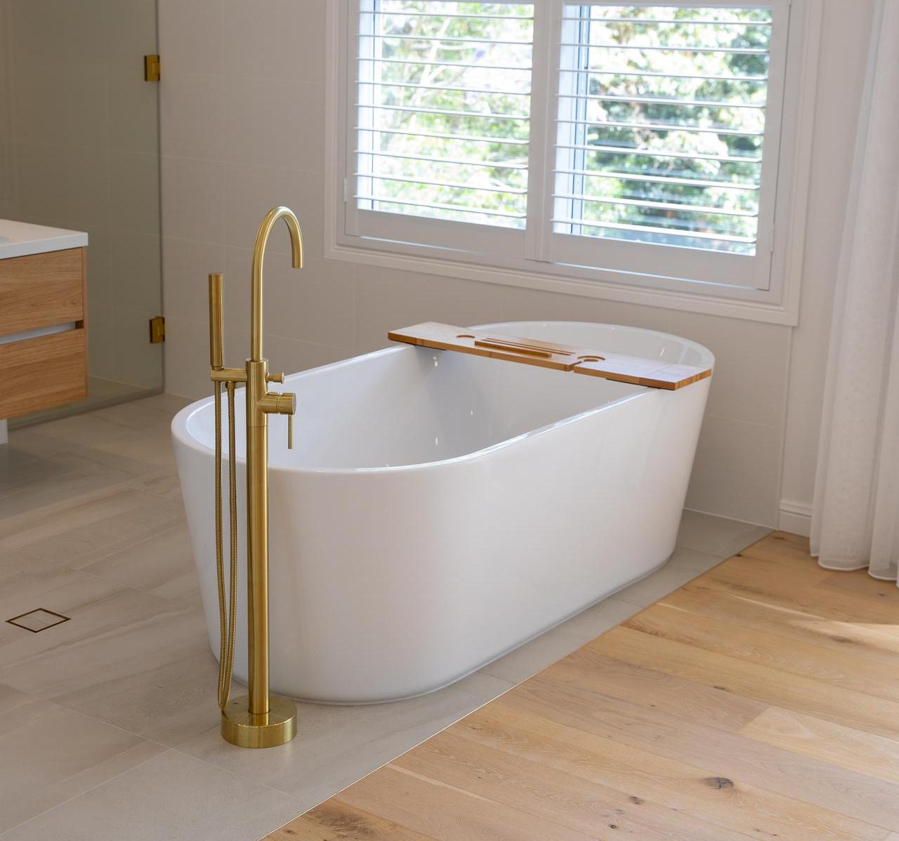 Modern bathroom with white freestanding tub, brass faucet, wood tray, and large window with shutters.