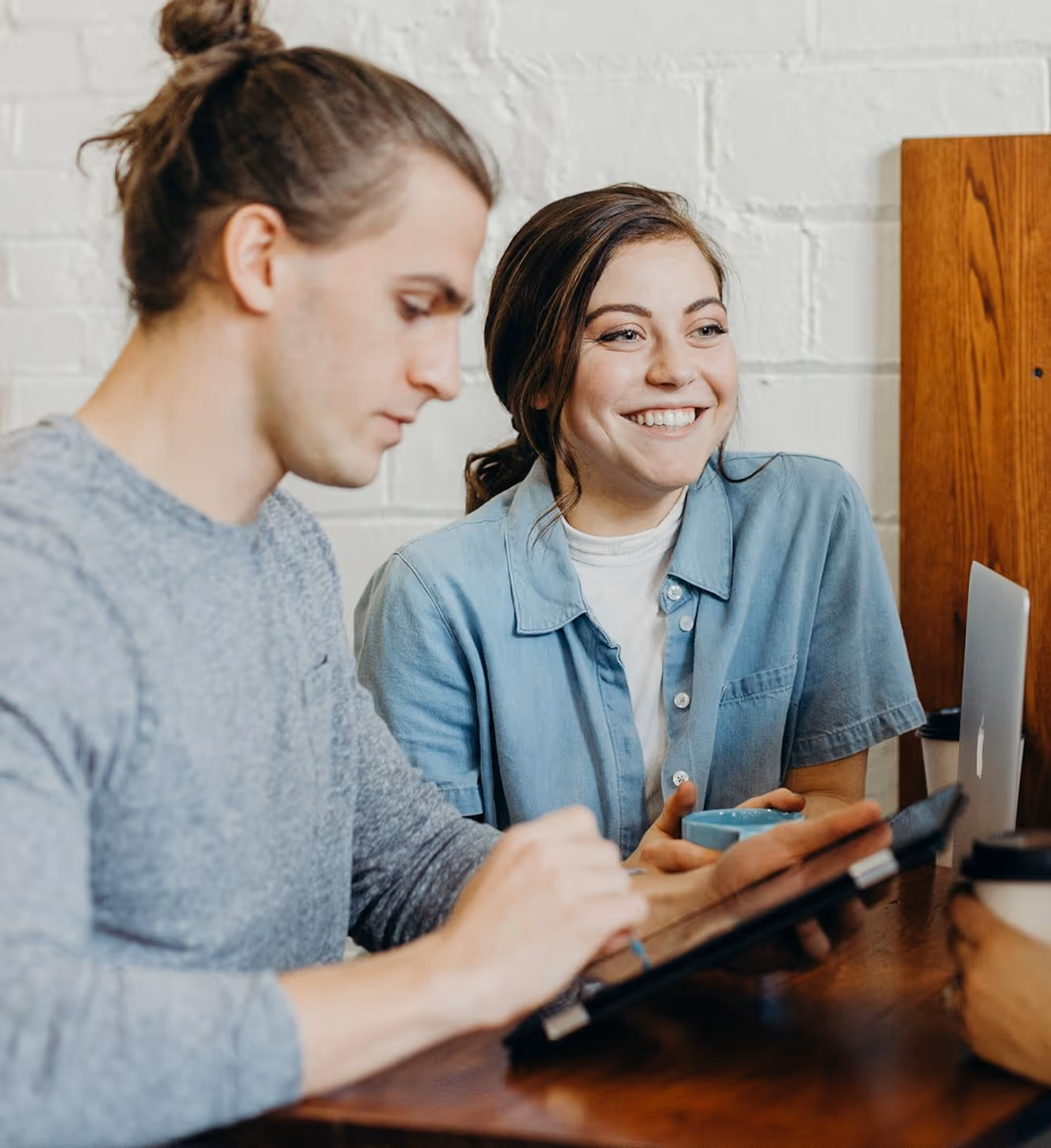Two people at a table, one using a tablet and the other smiling while holding a cup.