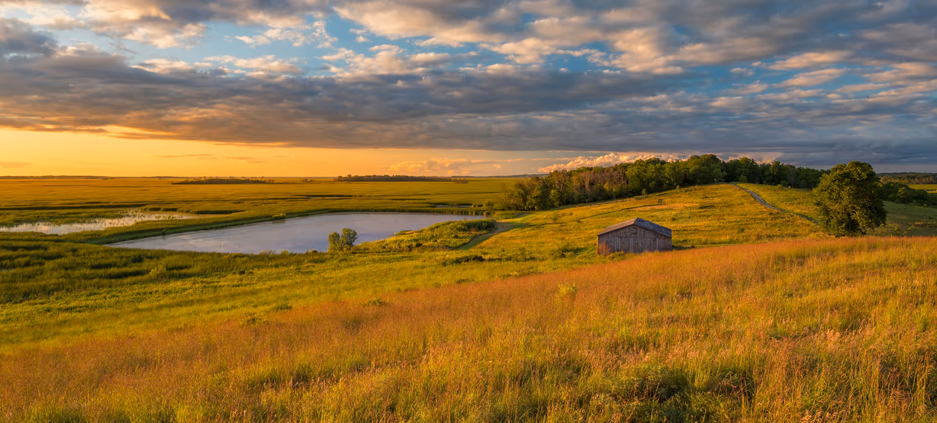 Sunset over grassy rolling hills with a small pond, trees, and a wooden barn.