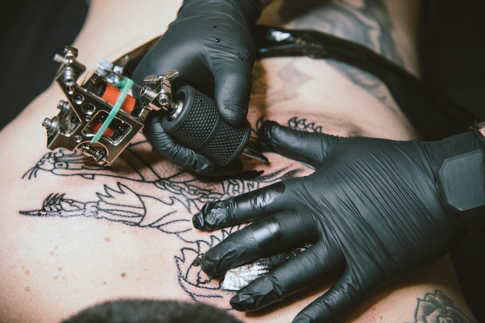 Tattoo artist wearing black gloves inks a large black line tattoo on a person's chest.