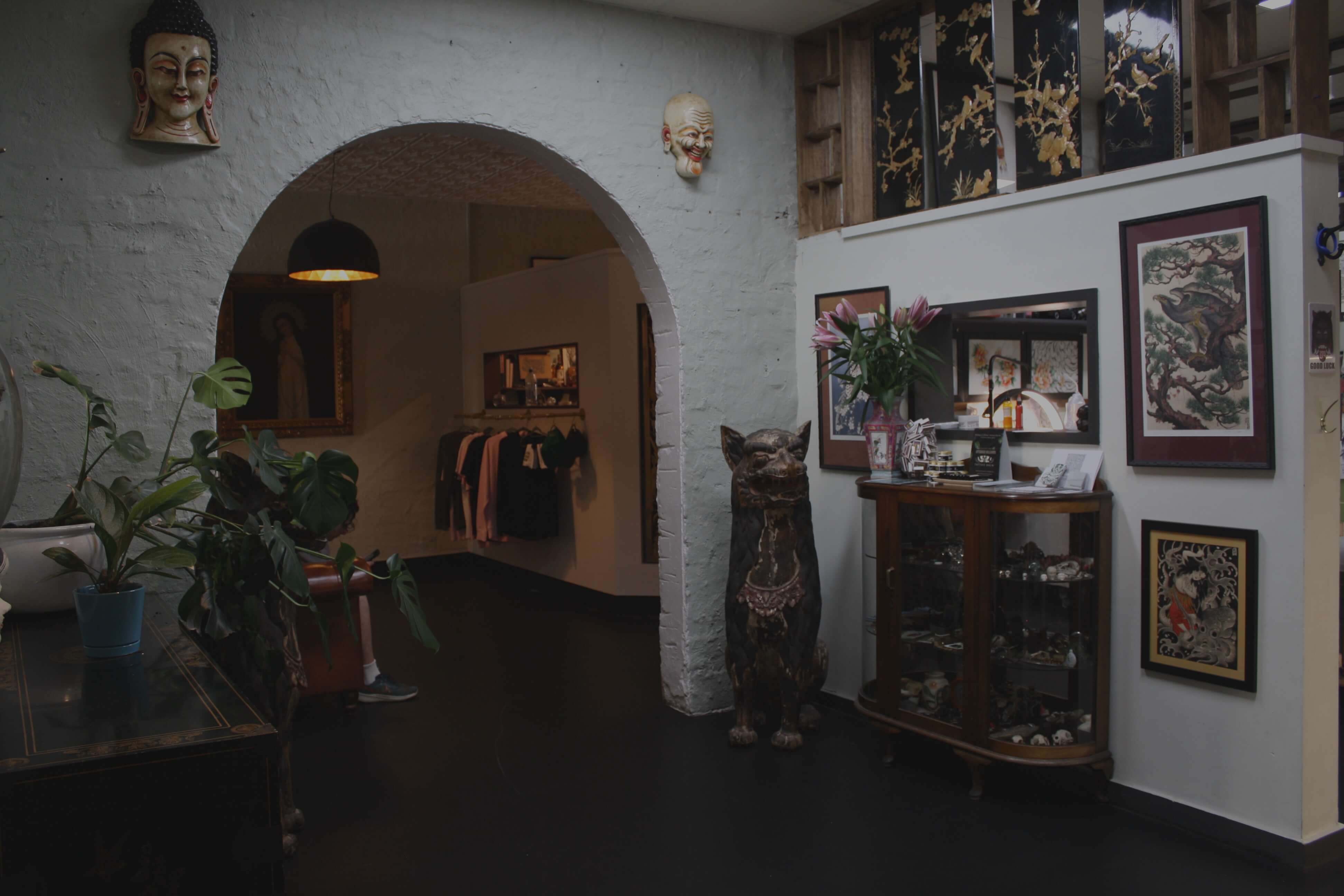 Interior room with plants, sculptures, framed artwork, and a white archway leading to a clothing display.