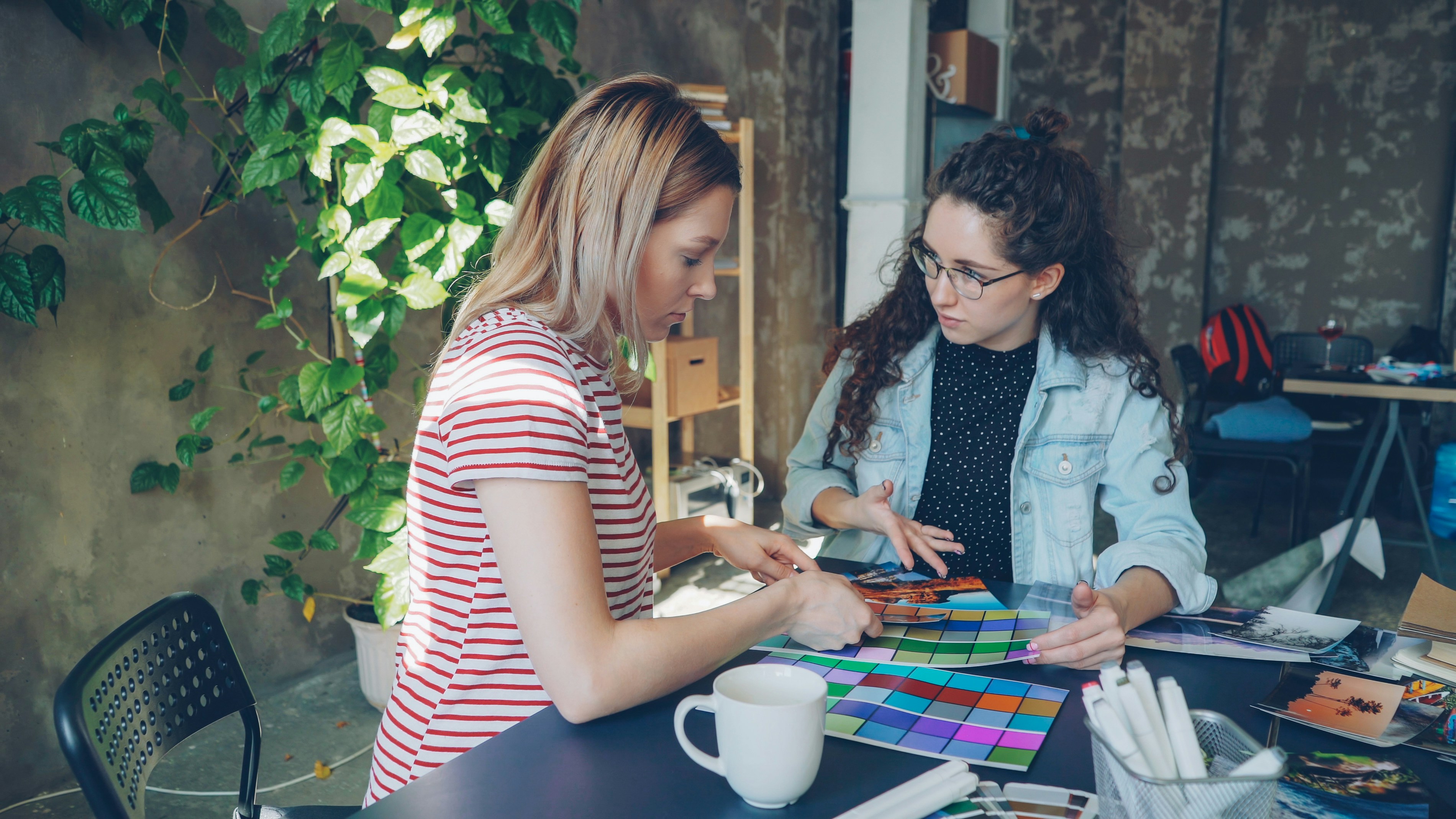 Two women examining color swatches and photos at a table in a casual workspace.