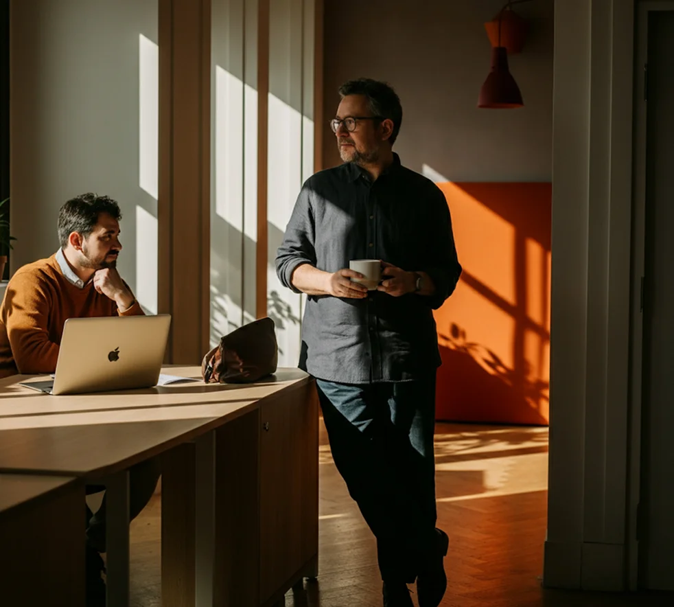 Man in black shirt holding cup standing by seated man with laptop in sunlit office.