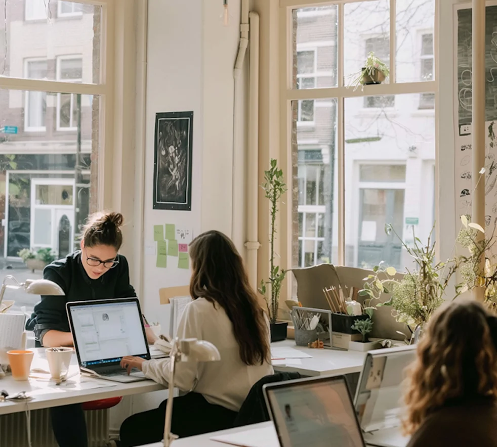 Three people working on laptops in a bright office space with large windows and plants.
