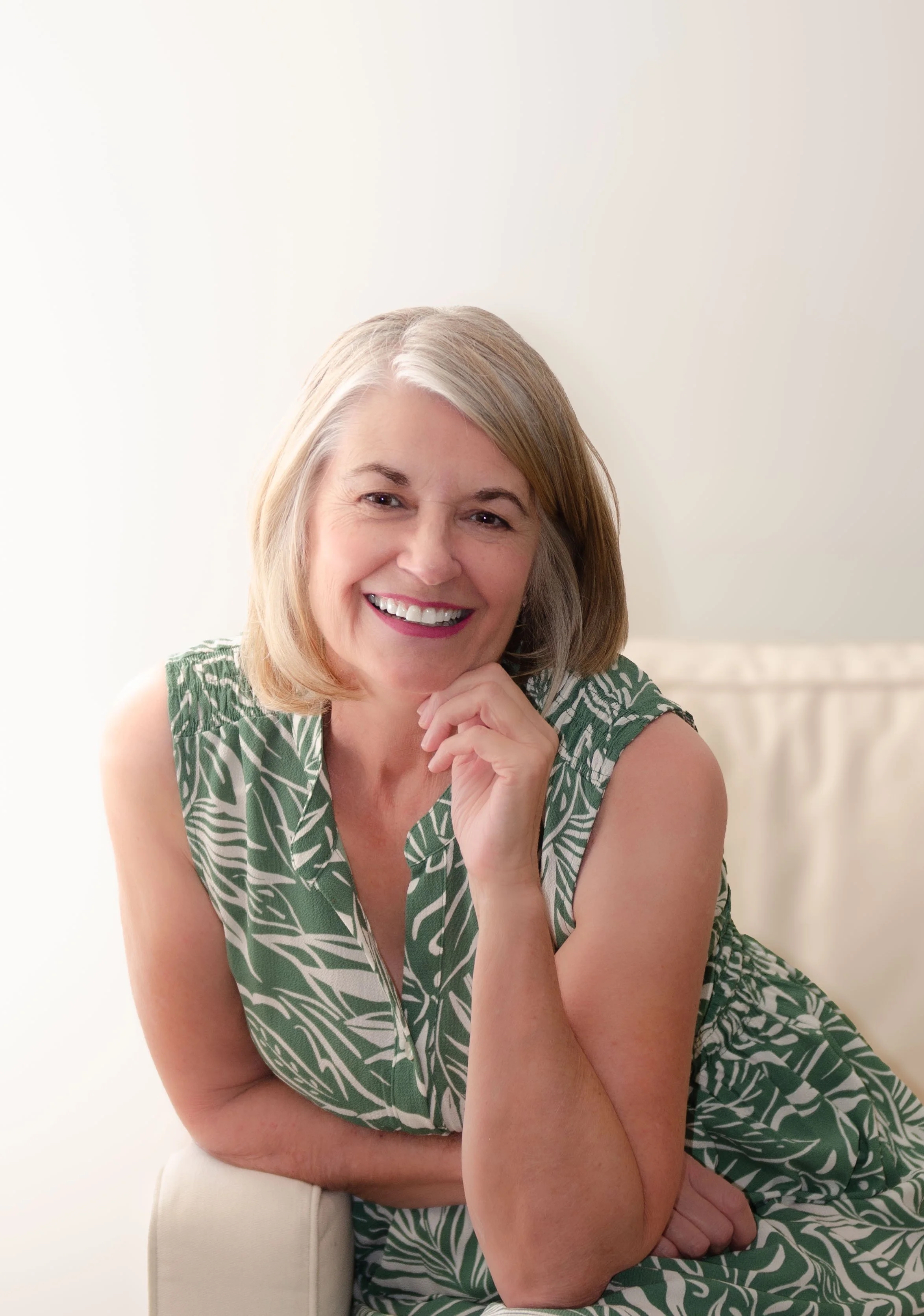Smiling older woman with short gray hair wearing a green patterned dress sitting on a white chair.