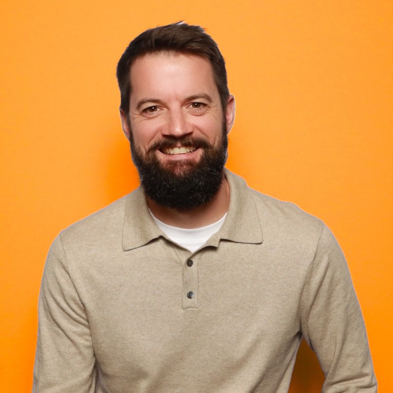 Smiling man with dark hair and beard wearing a beige sweater over a white shirt on orange background.