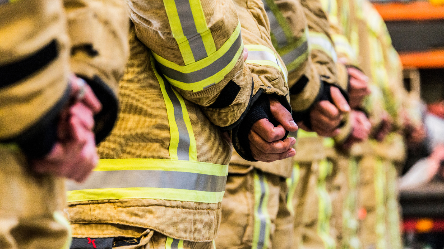 Close-up of firefighters standing side by side with hands clasped in front, wearing yellow protective gear with reflective stripes.