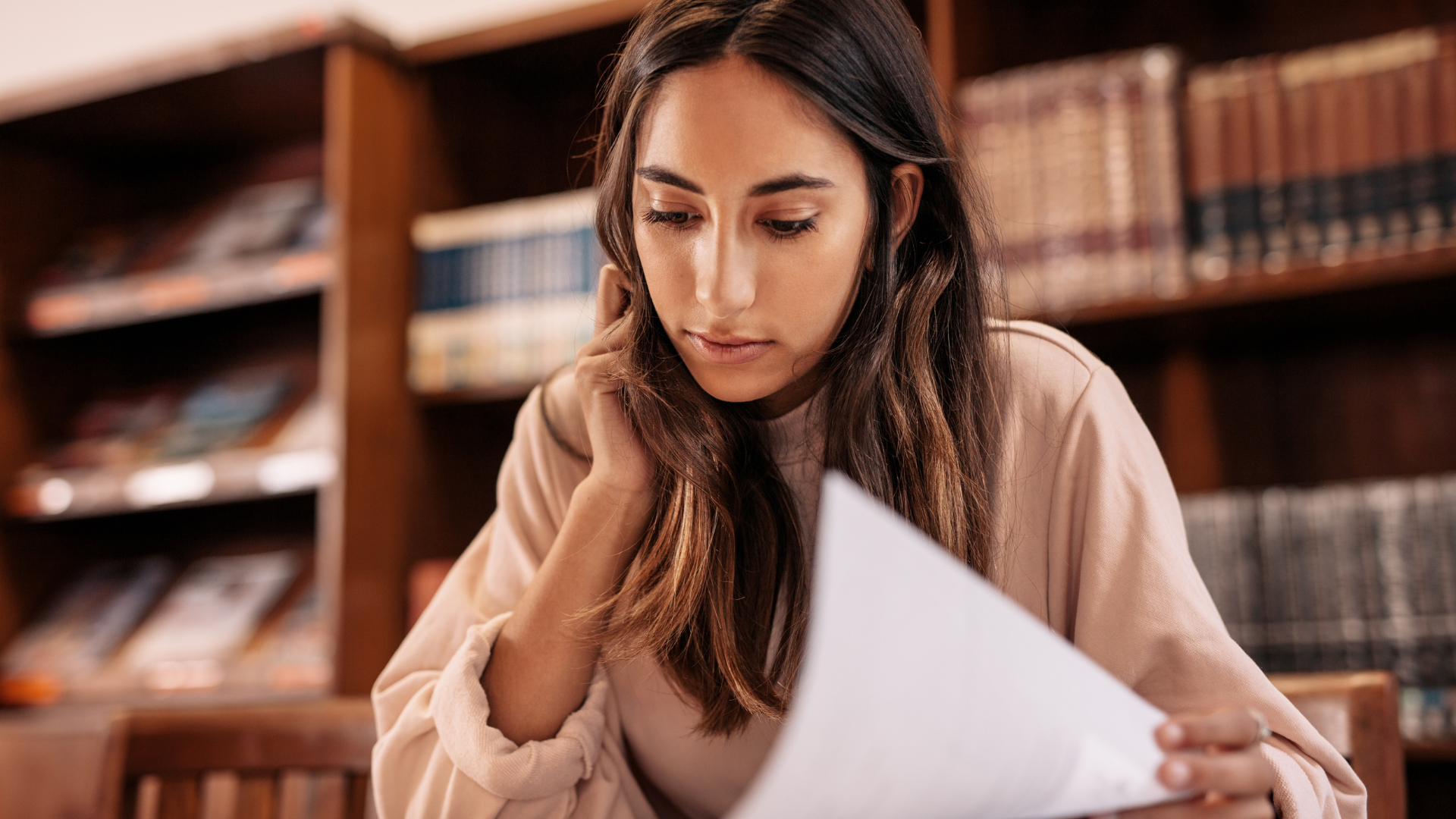 Young woman reading a document in a library with bookshelves in the background.