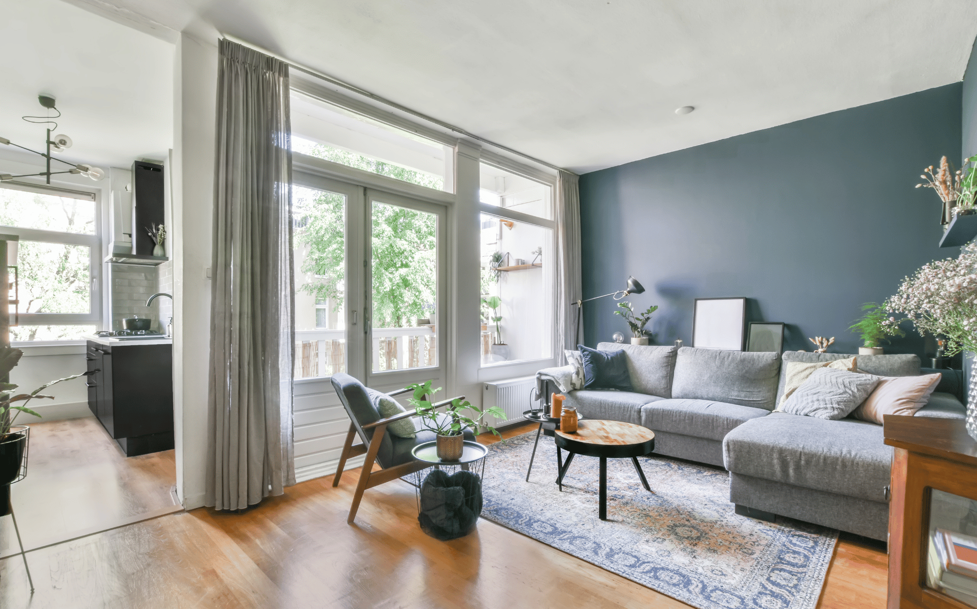 Helles Wohnzimmer mit dunkelblauer Akzentwand, großen Fenstern, Sofa und offener Küche.