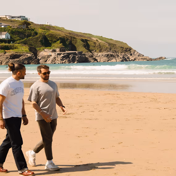 Two men walking side by side on a sandy beach with cliffs and ocean waves in the background.