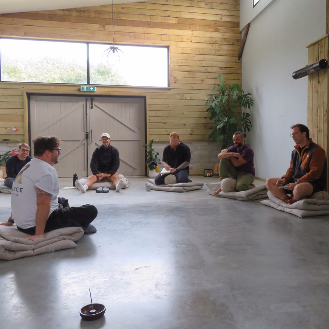 Six people sitting on cushions in a wooden room arranged in a circle for a group session.