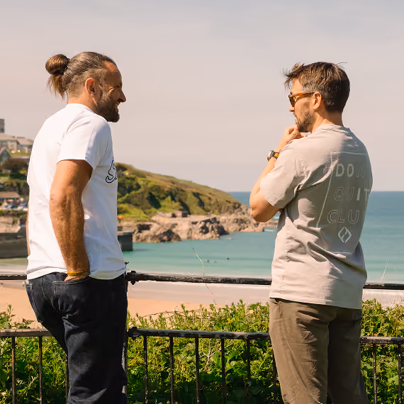 Two men talk near a railing overlooking a beach, ocean, and hills on a sunny day.