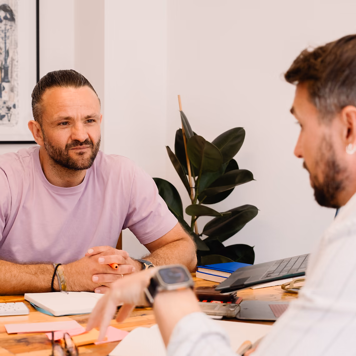 Two men at a desk in a discussion with notebooks, laptop, and plant in the background.