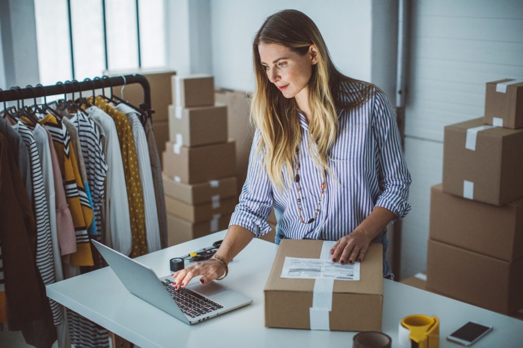 A woman in a striped shirt stands at a desk with her laptop and a package. Clothing on a rack and boxes are visible in the background, indicating an active workspace.