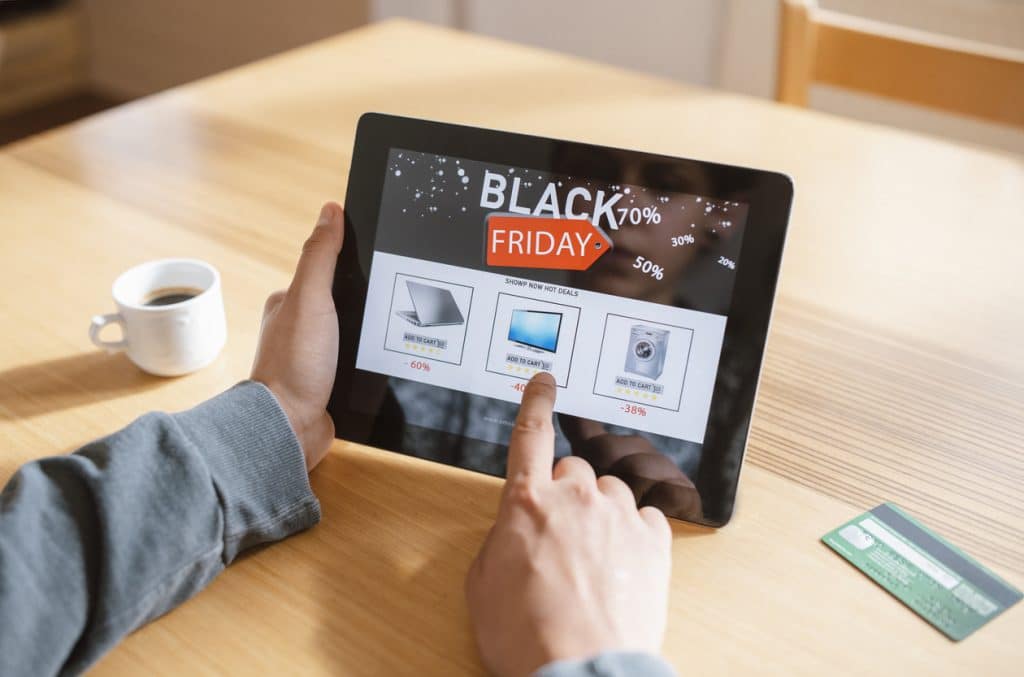 A person is browsing a tablet for online deals, with a credit card and coffee cup nearby. A screen displays a Shopify AI Concierge suggesting product options.