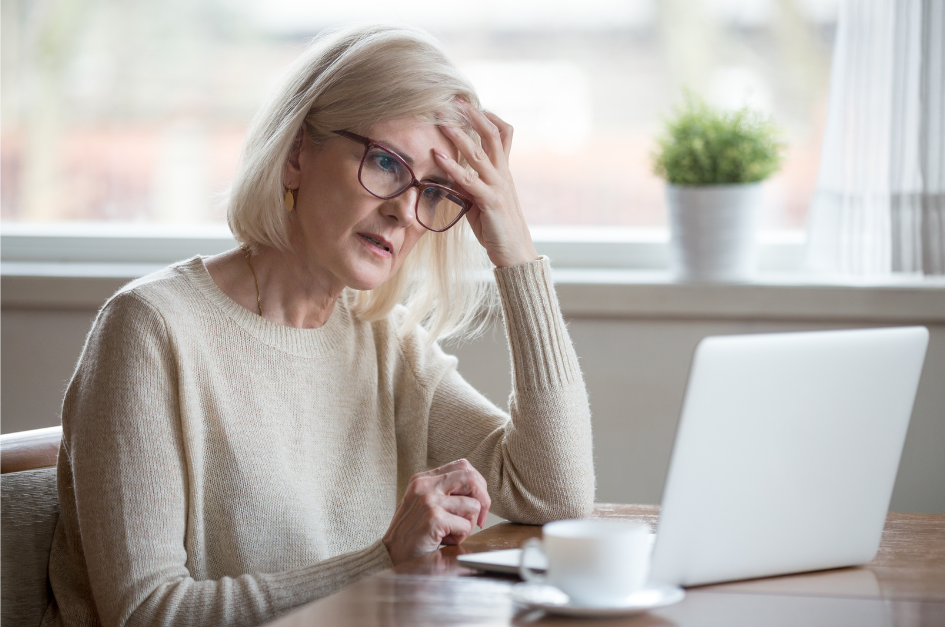 A concerned woman with glasses is intently looking at a laptop in a cozy room, surrounded by a warm cup of coffee and a small plant.