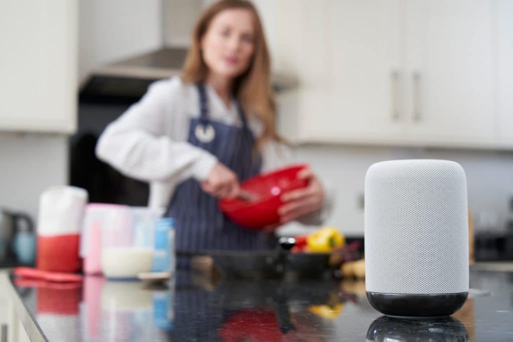 A smart speaker on a kitchen counter, with a blurred person in the background mixing ingredients in a bowl.