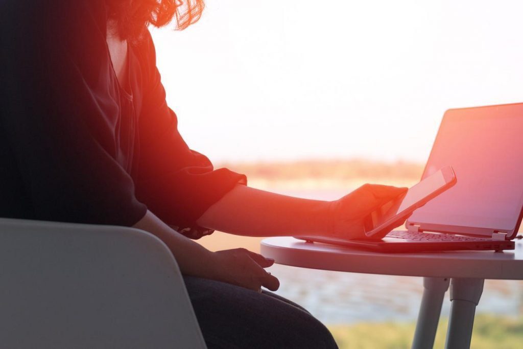 A person seated at a round table types on a smartphone next to an open laptop. Sunlight softly filters through the surroundings, casting a warm glow.