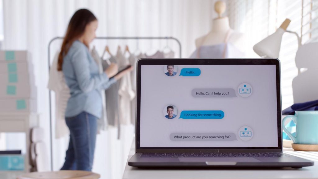 A laptop on a table shows a chatbot conversation with an AI Concierge. In the background, a woman browses through clothes on a rack.