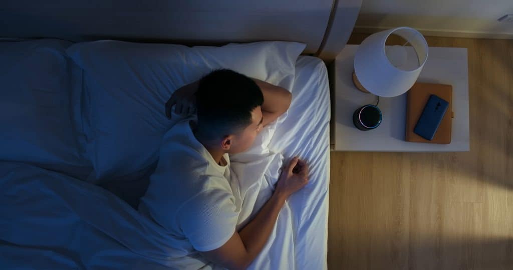 A person lies in bed, viewed from above. A lamp on the bedside table casts a warm glow over a closed notebook and a smartphone displaying notifications.
