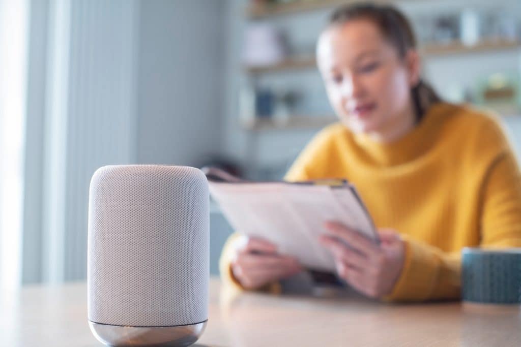 A smart speaker on a table with a softly blurred background showing a woman in a yellow sweater reading.